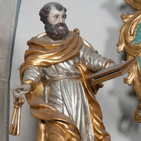 Zdjęcie nr 1: Rzeźba przedstawiająca św. Piotra, ścięta z tyłu, drążona, ustawiona na rzeźbionym fragmencie skały. Figura ukazana frontalnie z głową skierowaną delikatnie w prawo, obie ręce rozłożone w boki: w lewej trzyma zamkniętą księgę, a w prawej klucze. Twarz szeroka z otwartymi ustami, okolona gęstą brodą, włosy w postaci pukli po bokach i na czubku głowy. Święty ubrany jest w delikatnie drapowaną suknię, przepasaną w talii oraz płaszcz zapięty na lewym ramieniu, opadający na plecy i na prawe kolano, drapowany w grube fałdy, zawijające się przy brzegach; na stopach ma sandały. Polichromia naturalistyczna w odsłoniętych partiach ciała, szaty i atrybuty złocone oraz srebrzone.