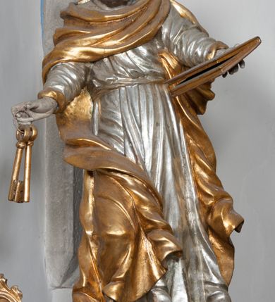 Zdjęcie nr 4: Rzeźba przedstawiająca św. Piotra, ścięta z tyłu, drążona, ustawiona na rzeźbionym fragmencie skały. Figura ukazana frontalnie z głową skierowaną delikatnie w prawo, obie ręce rozłożone w boki: w lewej trzyma zamkniętą księgę, a w prawej klucze. Twarz szeroka z otwartymi ustami, okolona gęstą brodą, włosy w postaci pukli po bokach i na czubku głowy. Święty ubrany jest w delikatnie drapowaną suknię, przepasaną w talii oraz płaszcz zapięty na lewym ramieniu, opadający na plecy i na prawe kolano, drapowany w grube fałdy, zawijające się przy brzegach; na stopach ma sandały. Polichromia naturalistyczna w odsłoniętych partiach ciała, szaty i atrybuty złocone oraz srebrzone.