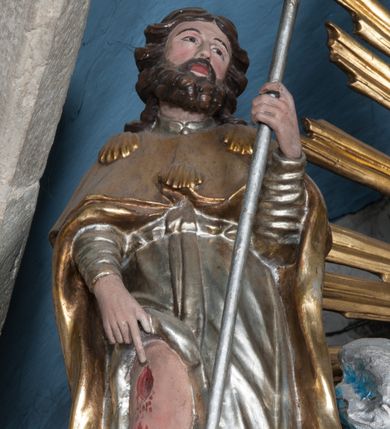 Zdjęcie nr 1: Rzeźba drążona, przedstawiająca św. Rocha. Figura ustawiona frontalnie, lewa ręka ugięta w łokciu, trzyma w niej kij pielgrzyma, prawa opuszczona w dół wskazuje na ranę na nodze. Twarz szeroka z małymi ustami, długim i prostym nosem, podłużnymi oczami okolonymi wąskimi łukami brwiowymi; broda krótka i gęsta; włosy sięgające do ramion, zawinięte do tyłu w bujne loki. Święty jest ubrany w srebrzoną suknię z długimi rękawami, odsłaniającą prawą nogę oraz złocony płaszcz pielgrzymi, zdobiony u góry trzema muszlami. U stóp świętego pies z kawałkiem chleba w pysku. Polichromia w odsłoniętych partiach ciała naturalistyczna, szaty złocone i srebrzone.