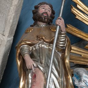 Zdjęcie nr 1: Rzeźba drążona, przedstawiająca św. Rocha. Figura ustawiona frontalnie, lewa ręka ugięta w łokciu, trzyma w niej kij pielgrzyma, prawa opuszczona w dół wskazuje na ranę na nodze. Twarz szeroka z małymi ustami, długim i prostym nosem, podłużnymi oczami okolonymi wąskimi łukami brwiowymi; broda krótka i gęsta; włosy sięgające do ramion, zawinięte do tyłu w bujne loki. Święty jest ubrany w srebrzoną suknię z długimi rękawami, odsłaniającą prawą nogę oraz złocony płaszcz pielgrzymi, zdobiony u góry trzema muszlami. U stóp świętego pies z kawałkiem chleba w pysku. Polichromia w odsłoniętych partiach ciała naturalistyczna, szaty złocone i srebrzone.