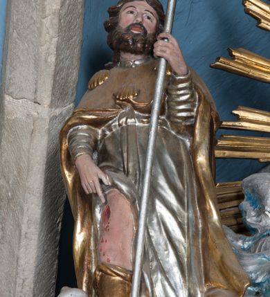 Zdjęcie nr 2: Rzeźba drążona, przedstawiająca św. Rocha. Figura ustawiona frontalnie, lewa ręka ugięta w łokciu, trzyma w niej kij pielgrzyma, prawa opuszczona w dół wskazuje na ranę na nodze. Twarz szeroka z małymi ustami, długim i prostym nosem, podłużnymi oczami okolonymi wąskimi łukami brwiowymi; broda krótka i gęsta; włosy sięgające do ramion, zawinięte do tyłu w bujne loki. Święty jest ubrany w srebrzoną suknię z długimi rękawami, odsłaniającą prawą nogę oraz złocony płaszcz pielgrzymi, zdobiony u góry trzema muszlami. U stóp świętego pies z kawałkiem chleba w pysku. Polichromia w odsłoniętych partiach ciała naturalistyczna, szaty złocone i srebrzone.