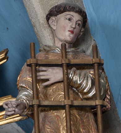 Zdjęcie nr 1: Rzeźba przedstawia św. Wawrzyńca. Święty ustawiony jest frontalnie z głową delikatnie zwróconą w swoją lewą stroną, z prawą ręką zgiętą w łokciu i wysuniętą do przodu, ułożoną do trzymania niezachowanego atrybutu (zapewne palmy męczeństwa); lewą ma złożoną na piersi, podtrzymuje nią duży ruszt w kształcie kratownicy. Twarz szeroka z małymi i delikatnie otwartymi ustami, z prostym nosem, z małymi oczami okolonymi wąskimi łukami brwiowymi. Włosy krótkie, kędzierzawe, ucięte w prostej linii wzdłuż nad uszami. Święty jest ubrany w długą, srebrzoną albę z długimi rękawami oraz złoconą dalmatykę, która zdobiona jest rzeźbioną wicią roślinną, na stopach ma złocone buty. Polichromia w odsłoniętych partiach ciała naturalistyczna, jasna, szaty złocone i srebrzone.