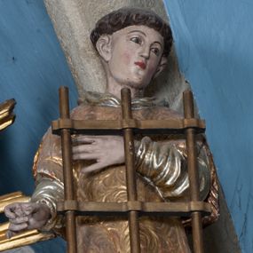 Zdjęcie nr 1: Rzeźba przedstawia św. Wawrzyńca. Święty ustawiony jest frontalnie z głową delikatnie zwróconą w swoją lewą stroną, z prawą ręką zgiętą w łokciu i wysuniętą do przodu, ułożoną do trzymania niezachowanego atrybutu (zapewne palmy męczeństwa); lewą ma złożoną na piersi, podtrzymuje nią duży ruszt w kształcie kratownicy. Twarz szeroka z małymi i delikatnie otwartymi ustami, z prostym nosem, z małymi oczami okolonymi wąskimi łukami brwiowymi. Włosy krótkie, kędzierzawe, ucięte w prostej linii wzdłuż nad uszami. Święty jest ubrany w długą, srebrzoną albę z długimi rękawami oraz złoconą dalmatykę, która zdobiona jest rzeźbioną wicią roślinną, na stopach ma złocone buty. Polichromia w odsłoniętych partiach ciała naturalistyczna, jasna, szaty złocone i srebrzone.