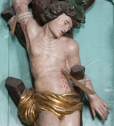Zdjęcie nr 1: Rzeźba ścięta z tyłu, przedstawia św. Sebastiana. Święty przywiązany do pnia w postawie stojącej z prawą nogą ugiętą w kolanie, z rękami i nogami skrępowanymi sznurami, głowa przechylona w jego lewą stronę, prawa ręka przywiązana do pnia wysoko nad głową. Twarz o młodzieńczych rysach, szeroka, usta drobne i otwarte, nos krótki i prosty, oczy podłużne okolone delikatną linią brwiową. Włosy ciemnobrązowe, średniej długości, zasłaniające uszy, zaczesane do tyłu bujnymi lokami. Ciało owinięte w biodrach złoconą przepaską. Święty ugodzony jedną strzałą w lewy bok, z którego tryska strumień krwi. Polichromia w odsłoniętych partiach ciała naturalistyczna, szaty i detale złocone.
