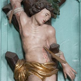Zdjęcie nr 1: Rzeźba ścięta z tyłu, przedstawia św. Sebastiana. Święty przywiązany do pnia w postawie stojącej z prawą nogą ugiętą w kolanie, z rękami i nogami skrępowanymi sznurami, głowa przechylona w jego lewą stronę, prawa ręka przywiązana do pnia wysoko nad głową. Twarz o młodzieńczych rysach, szeroka, usta drobne i otwarte, nos krótki i prosty, oczy podłużne okolone delikatną linią brwiową. Włosy ciemnobrązowe, średniej długości, zasłaniające uszy, zaczesane do tyłu bujnymi lokami. Ciało owinięte w biodrach złoconą przepaską. Święty ugodzony jedną strzałą w lewy bok, z którego tryska strumień krwi. Polichromia w odsłoniętych partiach ciała naturalistyczna, szaty i detale złocone.
