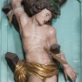 Zdjęcie nr 1: Rzeźba ścięta z tyłu, przedstawia św. Sebastiana. Święty przywiązany do pnia w postawie stojącej z prawą nogą ugiętą w kolanie, z rękami i nogami skrępowanymi sznurami, głowa przechylona w jego lewą stronę, prawa ręka przywiązana do pnia wysoko nad głową. Twarz o młodzieńczych rysach, szeroka, usta drobne i otwarte, nos krótki i prosty, oczy podłużne okolone delikatną linią brwiową. Włosy ciemnobrązowe, średniej długości, zasłaniające uszy, zaczesane do tyłu bujnymi lokami. Ciało owinięte w biodrach złoconą przepaską. Święty ugodzony jedną strzałą w lewy bok, z którego tryska strumień krwi. Polichromia w odsłoniętych partiach ciała naturalistyczna, szaty i detale złocone.
