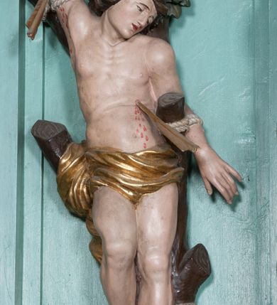 Zdjęcie nr 6: Rzeźba ścięta z tyłu, przedstawia św. Sebastiana. Święty przywiązany do pnia w postawie stojącej z prawą nogą ugiętą w kolanie, z rękami i nogami skrępowanymi sznurami, głowa przechylona w jego lewą stronę, prawa ręka przywiązana do pnia wysoko nad głową. Twarz o młodzieńczych rysach, szeroka, usta drobne i otwarte, nos krótki i prosty, oczy podłużne okolone delikatną linią brwiową. Włosy ciemnobrązowe, średniej długości, zasłaniające uszy, zaczesane do tyłu bujnymi lokami. Ciało owinięte w biodrach złoconą przepaską. Święty ugodzony jedną strzałą w lewy bok, z którego tryska strumień krwi. Polichromia w odsłoniętych partiach ciała naturalistyczna, szaty i detale złocone.
