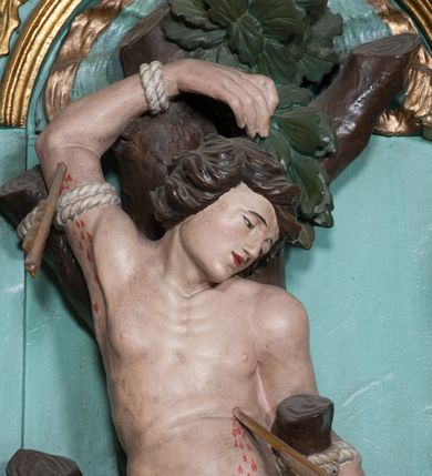 Zdjęcie nr 8: Rzeźba ścięta z tyłu, przedstawia św. Sebastiana. Święty przywiązany do pnia w postawie stojącej z prawą nogą ugiętą w kolanie, z rękami i nogami skrępowanymi sznurami, głowa przechylona w jego lewą stronę, prawa ręka przywiązana do pnia wysoko nad głową. Twarz o młodzieńczych rysach, szeroka, usta drobne i otwarte, nos krótki i prosty, oczy podłużne okolone delikatną linią brwiową. Włosy ciemnobrązowe, średniej długości, zasłaniające uszy, zaczesane do tyłu bujnymi lokami. Ciało owinięte w biodrach złoconą przepaską. Święty ugodzony jedną strzałą w lewy bok, z którego tryska strumień krwi. Polichromia w odsłoniętych partiach ciała naturalistyczna, szaty i detale złocone.
