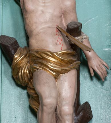 Zdjęcie nr 13: Rzeźba ścięta z tyłu, przedstawia św. Sebastiana. Święty przywiązany do pnia w postawie stojącej z prawą nogą ugiętą w kolanie, z rękami i nogami skrępowanymi sznurami, głowa przechylona w jego lewą stronę, prawa ręka przywiązana do pnia wysoko nad głową. Twarz o młodzieńczych rysach, szeroka, usta drobne i otwarte, nos krótki i prosty, oczy podłużne okolone delikatną linią brwiową. Włosy ciemnobrązowe, średniej długości, zasłaniające uszy, zaczesane do tyłu bujnymi lokami. Ciało owinięte w biodrach złoconą przepaską. Święty ugodzony jedną strzałą w lewy bok, z którego tryska strumień krwi. Polichromia w odsłoniętych partiach ciała naturalistyczna, szaty i detale złocone.
