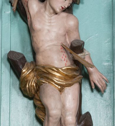 Zdjęcie nr 7: Rzeźba ścięta z tyłu, przedstawia św. Sebastiana. Święty przywiązany do pnia w postawie stojącej z prawą nogą ugiętą w kolanie, z rękami i nogami skrępowanymi sznurami, głowa przechylona w jego lewą stronę, prawa ręka przywiązana do pnia wysoko nad głową. Twarz o młodzieńczych rysach, szeroka, usta drobne i otwarte, nos krótki i prosty, oczy podłużne okolone delikatną linią brwiową. Włosy ciemnobrązowe, średniej długości, zasłaniające uszy, zaczesane do tyłu bujnymi lokami. Ciało owinięte w biodrach złoconą przepaską. Święty ugodzony jedną strzałą w lewy bok, z którego tryska strumień krwi. Polichromia w odsłoniętych partiach ciała naturalistyczna, szaty i detale złocone.
