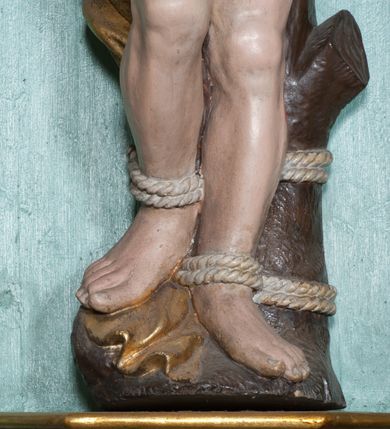 Zdjęcie nr 5: Rzeźba ścięta z tyłu, przedstawia św. Sebastiana. Święty przywiązany do pnia w postawie stojącej z prawą nogą ugiętą w kolanie, z rękami i nogami skrępowanymi sznurami, głowa przechylona w jego lewą stronę, prawa ręka przywiązana do pnia wysoko nad głową. Twarz o młodzieńczych rysach, szeroka, usta drobne i otwarte, nos krótki i prosty, oczy podłużne okolone delikatną linią brwiową. Włosy ciemnobrązowe, średniej długości, zasłaniające uszy, zaczesane do tyłu bujnymi lokami. Ciało owinięte w biodrach złoconą przepaską. Święty ugodzony jedną strzałą w lewy bok, z którego tryska strumień krwi. Polichromia w odsłoniętych partiach ciała naturalistyczna, szaty i detale złocone.
