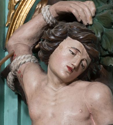 Zdjęcie nr 10: Rzeźba ścięta z tyłu, przedstawia św. Sebastiana. Święty przywiązany do pnia w postawie stojącej z prawą nogą ugiętą w kolanie, z rękami i nogami skrępowanymi sznurami, głowa przechylona w jego lewą stronę, prawa ręka przywiązana do pnia wysoko nad głową. Twarz o młodzieńczych rysach, szeroka, usta drobne i otwarte, nos krótki i prosty, oczy podłużne okolone delikatną linią brwiową. Włosy ciemnobrązowe, średniej długości, zasłaniające uszy, zaczesane do tyłu bujnymi lokami. Ciało owinięte w biodrach złoconą przepaską. Święty ugodzony jedną strzałą w lewy bok, z którego tryska strumień krwi. Polichromia w odsłoniętych partiach ciała naturalistyczna, szaty i detale złocone.

