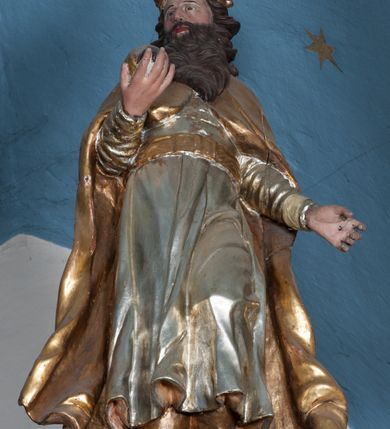 Zdjęcie nr 2: Rzeźba ścięta z tyłu, drążona, przedstawia św. Stefana. Figura ustawiona frontalnie w delikatnym kontrapoście z lewą nogą ugiętą w kolanie, z głową skierowaną w prawo. W prawej dłoni trzyma jabłko królewskie, lewą ma odsuniętą w bok z dłonią ułożoną do trzymania niezachowanego atrybutu (berła). Twarz szeroka, okolona ciemną, bujną i rozwianą brodą. Włosy ciemne, średniej długości, zasłaniające uszy i zawinięte dużymi lokami do tyłu. Święty ubrany jest w buty z wysokimi cholewami, srebrzoną suknię z długimi rękawami, przewiązaną pasem oraz złocony płaszcz, spięty na piersiach o dekoracyjnie wywiniętych fałdach. Na głowie zamknięta korona. Polichromia naturalistyczna w odsłoniętych partiach ciała, strój i atrybuty złocone.




