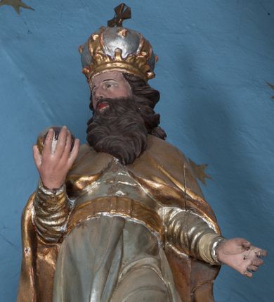 Zdjęcie nr 1: Rzeźba ścięta z tyłu, drążona, przedstawia św. Stefana. Figura ustawiona frontalnie w delikatnym kontrapoście z lewą nogą ugiętą w kolanie, z głową skierowaną w prawo. W prawej dłoni trzyma jabłko królewskie, lewą ma odsuniętą w bok z dłonią ułożoną do trzymania niezachowanego atrybutu (berła). Twarz szeroka, okolona ciemną, bujną i rozwianą brodą. Włosy ciemne, średniej długości, zasłaniające uszy i zawinięte dużymi lokami do tyłu. Święty ubrany jest w buty z wysokimi cholewami, srebrzoną suknię z długimi rękawami, przewiązaną pasem oraz złocony płaszcz, spięty na piersiach o dekoracyjnie wywiniętych fałdach. Na głowie zamknięta korona. Polichromia naturalistyczna w odsłoniętych partiach ciała, strój i atrybuty złocone.




