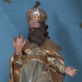 Zdjęcie nr 1: Rzeźba ścięta z tyłu, drążona, przedstawia św. Stefana. Figura ustawiona frontalnie w delikatnym kontrapoście z lewą nogą ugiętą w kolanie, z głową skierowaną w prawo. W prawej dłoni trzyma jabłko królewskie, lewą ma odsuniętą w bok z dłonią ułożoną do trzymania niezachowanego atrybutu (berła). Twarz szeroka, okolona ciemną, bujną i rozwianą brodą. Włosy ciemne, średniej długości, zasłaniające uszy i zawinięte dużymi lokami do tyłu. Święty ubrany jest w buty z wysokimi cholewami, srebrzoną suknię z długimi rękawami, przewiązaną pasem oraz złocony płaszcz, spięty na piersiach o dekoracyjnie wywiniętych fałdach. Na głowie zamknięta korona. Polichromia naturalistyczna w odsłoniętych partiach ciała, strój i atrybuty złocone.





