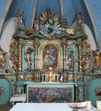 Zdjęcie nr 24: Nastawa ołtarzowa architektoniczna, jednokondygnacyjna, trójosiowa ze zwieńczeniem i z bramkami. Ołtarz prostopadłościenny, drewniany, mensa drewniana z kamiennym portatylem. Antependium w formie leżącego prostokąta ujętego złoconą, profilowaną ramą. W polu obraz malowany olejem na płótnie. W centrum kartusz w kształcie wydłużonego czwórliścia, dekorowany muszlą i wolutami z przedstawieniem pejzażu i widokiem na Golgotę. Po bokach kartusza bujne gałązki kwiatów. Tabernakulum w formie prostopadłościennej skrzynki o wypukłej ścianie frontowej, na profilowanym cokole, zamknięte profilowanym gzymsem, ujęte uszami utworzonymi z liści. Nad drzwiczkami, na gzymsie kartusz z niewielkim krucyfiksem. Powyżej tron wystawienia w formie profilowanej podstawy dekorowanej liśćmi. Na ściance frontowej tabernakulum profilowana rama w kształcie stojącego prostokąta zamkniętego łukiem nadwieszonym, w polu drzwiczki w formie stojącego prostokąta z przedstawieniem kielicha w glorii, powyżej drzwiczek winne grono. 

Nastawa ustawiona na zdwojonym cokole, dekorowanym wijącym się akantem. Osie kondygnacji wydzielone dwiema parami pilastrów oraz ustawionymi ukośnie kolumnami. Podpory dźwigają pełne belkowanie, przerwane nad polem środkowym i wyłamane nad osiami bocznymi, na których skraju ustawiony jest ukośnie przerwany, wolutowy przyczółek. Kolumny o kręconych trzonach, owinięte wicią liści akantu, kapitele korynckie; imposty ustawione pod kątem. W polu środkowym rama w kształcie stojącego prostokąta zamkniętego łukiem półkolistym z obrazem św. Kwiryna. W osiach bocznych płytkie płyciny, zwieńczone konchą z figurami św. Sebastiana po lewej stronie i św. Jana Nepomucena po prawej stronie, ustawionymi na wspornikach. Kondygnacja ujęta jest po bokach szerokimi uszami utworzonymi z liści akantu i karbowanej wstęgi. Fryz nad osiami bocznymi zdobiony kartuszami, we fryzie impostów z dwóch stron rozety; przyłucza osi bocznych dekorowane akantem.

W zwieńczeniu rzeźbiona półpostać Boga Ojca na obłoku z widocznym poniżej fragmentem globu ziemskiego i gołębicą Ducha Świętego; całość ukazana na tle promienistej glorii. Na belkowaniu, na osiach podpór wewnętrznych ustawione są rzeźby: św. Rocha  po lewej stronie i św. Wawrzyńca po prawej, a na zewnętrznych osiach retabulum rzeźby: Karola Boromeusza po lewej stronie i św. Stefana po prawej.

Nastawa ołtarzowa ujęta jest po bokach szerokimi bramkami w kształcie stojących prostokątów z drzwiczkami zamkniętymi półkoliście. Bramki ujęte są pilastrami o kanelowanych trzonach i fantazyjnych kapitelach, wspierających niepełne belkowanie oraz przerwany przyczółek. Nad bramkami ustawione są na cokołach rzeźby: św. Piotra po lewej stronie i św. Pawła po prawej. W polu drzwiczek owalne kartusze z herbem Görgey. W prawym kartuszu opisanym słowami: „BALTHAZAR GÖRGEI SENIOR”. Nad arkadami bramek są muszle, w przyłuczach akant, we fryzie rozety, a przerwany przyczółek dekorowany jest akantem. Struktura w całości polichromowana w odcieniu bladozielonym; ornamentyka, kolumny i profilowania złocone, detale srebrzone.