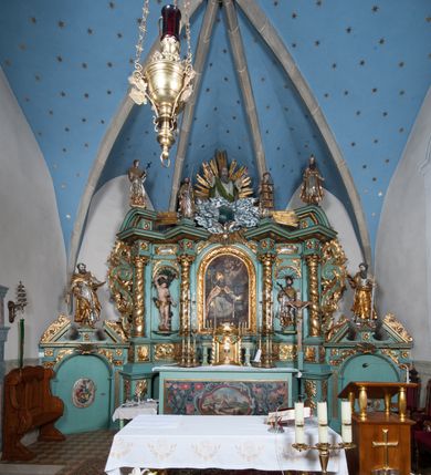 Zdjęcie nr 13: Nastawa ołtarzowa architektoniczna, jednokondygnacyjna, trójosiowa ze zwieńczeniem i z bramkami. Ołtarz prostopadłościenny, drewniany, mensa drewniana z kamiennym portatylem. Antependium w formie leżącego prostokąta ujętego złoconą, profilowaną ramą. W polu obraz malowany olejem na płótnie. W centrum kartusz w kształcie wydłużonego czwórliścia, dekorowany muszlą i wolutami z przedstawieniem pejzażu i widokiem na Golgotę. Po bokach kartusza bujne gałązki kwiatów. Tabernakulum w formie prostopadłościennej skrzynki o wypukłej ścianie frontowej, na profilowanym cokole, zamknięte profilowanym gzymsem, ujęte uszami utworzonymi z liści. Nad drzwiczkami, na gzymsie kartusz z niewielkim krucyfiksem. Powyżej tron wystawienia w formie profilowanej podstawy dekorowanej liśćmi. Na ściance frontowej tabernakulum profilowana rama w kształcie stojącego prostokąta zamkniętego łukiem nadwieszonym, w polu drzwiczki w formie stojącego prostokąta z przedstawieniem kielicha w glorii, powyżej drzwiczek winne grono. 

Nastawa ustawiona na zdwojonym cokole, dekorowanym wijącym się akantem. Osie kondygnacji wydzielone dwiema parami pilastrów oraz ustawionymi ukośnie kolumnami. Podpory dźwigają pełne belkowanie, przerwane nad polem środkowym i wyłamane nad osiami bocznymi, na których skraju ustawiony jest ukośnie przerwany, wolutowy przyczółek. Kolumny o kręconych trzonach, owinięte wicią liści akantu, kapitele korynckie; imposty ustawione pod kątem. W polu środkowym rama w kształcie stojącego prostokąta zamkniętego łukiem półkolistym z obrazem św. Kwiryna. W osiach bocznych płytkie płyciny, zwieńczone konchą z figurami św. Sebastiana po lewej stronie i św. Jana Nepomucena po prawej stronie, ustawionymi na wspornikach. Kondygnacja ujęta jest po bokach szerokimi uszami utworzonymi z liści akantu i karbowanej wstęgi. Fryz nad osiami bocznymi zdobiony kartuszami, we fryzie impostów z dwóch stron rozety; przyłucza osi bocznych dekorowane akantem.

W zwieńczeniu rzeźbiona półpostać Boga Ojca na obłoku z widocznym poniżej fragmentem globu ziemskiego i gołębicą Ducha Świętego; całość ukazana na tle promienistej glorii. Na belkowaniu, na osiach podpór wewnętrznych ustawione są rzeźby: św. Rocha  po lewej stronie i św. Wawrzyńca po prawej, a na zewnętrznych osiach retabulum rzeźby: Karola Boromeusza po lewej stronie i św. Stefana po prawej.

Nastawa ołtarzowa ujęta jest po bokach szerokimi bramkami w kształcie stojących prostokątów z drzwiczkami zamkniętymi półkoliście. Bramki ujęte są pilastrami o kanelowanych trzonach i fantazyjnych kapitelach, wspierających niepełne belkowanie oraz przerwany przyczółek. Nad bramkami ustawione są na cokołach rzeźby: św. Piotra po lewej stronie i św. Pawła po prawej. W polu drzwiczek owalne kartusze z herbem Görgey. W prawym kartuszu opisanym słowami: „BALTHAZAR GÖRGEI SENIOR”. Nad arkadami bramek są muszle, w przyłuczach akant, we fryzie rozety, a przerwany przyczółek dekorowany jest akantem. Struktura w całości polichromowana w odcieniu bladozielonym; ornamentyka, kolumny i profilowania złocone, detale srebrzone.