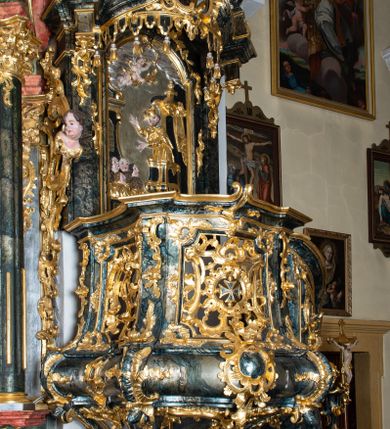 Zdjęcie nr 1: Ambona przyścienna, wisząca, ustawiona przy północnym filarze łuku tęczowego; dostępna jednobiegowymi schodami z balustradą od strony prezbiterium. Kosz na planie czworoboku, u dołu wydzielony silnie występującym ćwierćwałkiem z nałożoną złoconą dekoracją rocaille'ową; u góry zamknięty profilowanym gzymsem. Podwieszenie w formie odwróconego stożka. Ścianki kosza artykułowane pilastrami w kształcie spływów wolutowych. W polach prostokątne płyciny o wklęsłych narożach ozdobione bogato ażurowym ornamentem rokokowym. Zaplecek flankowany pilastrami podtrzymującymi przerwane belkowanie, które nad polem środkowym przechodzi w baldachim. W polu zaplecka prostokątna płycina zamknięta łukiem nadwieszonym z płaskorzeźbą św. Jana Nepomucena. Baldachim krótki, na rzucie półkolistym bez podniebienia, od dołu zdobiony lambrekinem. Na impostach cokoły utworzone z wolut, a na nich ustawione płomieniste wazony. Zwieńczenie w kształcie zbliżonym do kwadratu, ujętym z każdej strony spływami wolutowymi. W polu głównym owalna płycina, na której tle jest promienista gloria z gołębicą Ducha Świętego. Balustrada schodów pełna; na końcu zakończona filarkiem z rokokowym wazonem płomienistym. Całość silnie dekorowana ornamentem rocaille’owym. Struktura marmoryzowana w kolorze ciemnozielonym, ornamentyka i detale złocone oraz srebrzone.
