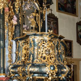 Zdjęcie nr 1: Ambona przyścienna, wisząca, ustawiona przy północnym filarze łuku tęczowego; dostępna jednobiegowymi schodami z balustradą od strony prezbiterium. Kosz na planie czworoboku, u dołu wydzielony silnie występującym ćwierćwałkiem z nałożoną złoconą dekoracją rocaille'ową; u góry zamknięty profilowanym gzymsem. Podwieszenie w formie odwróconego stożka. Ścianki kosza artykułowane pilastrami w kształcie spływów wolutowych. W polach prostokątne płyciny o wklęsłych narożach ozdobione bogato ażurowym ornamentem rokokowym. Zaplecek flankowany pilastrami podtrzymującymi przerwane belkowanie, które nad polem środkowym przechodzi w baldachim. W polu zaplecka prostokątna płycina zamknięta łukiem nadwieszonym z płaskorzeźbą św. Jana Nepomucena. Baldachim krótki, na rzucie półkolistym bez podniebienia, od dołu zdobiony lambrekinem. Na impostach cokoły utworzone z wolut, a na nich ustawione płomieniste wazony. Zwieńczenie w kształcie zbliżonym do kwadratu, ujętym z każdej strony spływami wolutowymi. W polu głównym owalna płycina, na której tle jest promienista gloria z gołębicą Ducha Świętego. Balustrada schodów pełna; na końcu zakończona filarkiem z rokokowym wazonem płomienistym. Całość silnie dekorowana ornamentem rocaille’owym. Struktura marmoryzowana w kolorze ciemnozielonym, ornamentyka i detale złocone oraz srebrzone.

