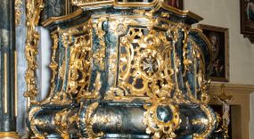 Zdjęcie nr 1: Ambona przyścienna, wisząca, ustawiona przy północnym filarze łuku tęczowego; dostępna jednobiegowymi schodami z balustradą od strony prezbiterium. Kosz na planie czworoboku, u dołu wydzielony silnie występującym ćwierćwałkiem z nałożoną złoconą dekoracją rocaille'ową; u góry zamknięty profilowanym gzymsem. Podwieszenie w formie odwróconego stożka. Ścianki kosza artykułowane pilastrami w kształcie spływów wolutowych. W polach prostokątne płyciny o wklęsłych narożach ozdobione bogato ażurowym ornamentem rokokowym. Zaplecek flankowany pilastrami podtrzymującymi przerwane belkowanie, które nad polem środkowym przechodzi w baldachim. W polu zaplecka prostokątna płycina zamknięta łukiem nadwieszonym z płaskorzeźbą św. Jana Nepomucena. Baldachim krótki, na rzucie półkolistym bez podniebienia, od dołu zdobiony lambrekinem. Na impostach cokoły utworzone z wolut, a na nich ustawione płomieniste wazony. Zwieńczenie w kształcie zbliżonym do kwadratu, ujętym z każdej strony spływami wolutowymi. W polu głównym owalna płycina, na której tle jest promienista gloria z gołębicą Ducha Świętego. Balustrada schodów pełna; na końcu zakończona filarkiem z rokokowym wazonem płomienistym. Całość silnie dekorowana ornamentem rocaille’owym. Struktura marmoryzowana w kolorze ciemnozielonym, ornamentyka i detale złocone oraz srebrzone.
