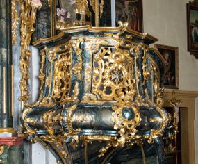 Zdjęcie nr 1: Ambona przyścienna, wisząca, ustawiona przy północnym filarze łuku tęczowego; dostępna jednobiegowymi schodami z balustradą od strony prezbiterium. Kosz na planie czworoboku, u dołu wydzielony silnie występującym ćwierćwałkiem z nałożoną złoconą dekoracją rocaille'ową; u góry zamknięty profilowanym gzymsem. Podwieszenie w formie odwróconego stożka. Ścianki kosza artykułowane pilastrami w kształcie spływów wolutowych. W polach prostokątne płyciny o wklęsłych narożach ozdobione bogato ażurowym ornamentem rokokowym. Zaplecek flankowany pilastrami podtrzymującymi przerwane belkowanie, które nad polem środkowym przechodzi w baldachim. W polu zaplecka prostokątna płycina zamknięta łukiem nadwieszonym z płaskorzeźbą św. Jana Nepomucena. Baldachim krótki, na rzucie półkolistym bez podniebienia, od dołu zdobiony lambrekinem. Na impostach cokoły utworzone z wolut, a na nich ustawione płomieniste wazony. Zwieńczenie w kształcie zbliżonym do kwadratu, ujętym z każdej strony spływami wolutowymi. W polu głównym owalna płycina, na której tle jest promienista gloria z gołębicą Ducha Świętego. Balustrada schodów pełna; na końcu zakończona filarkiem z rokokowym wazonem płomienistym. Całość silnie dekorowana ornamentem rocaille’owym. Struktura marmoryzowana w kolorze ciemnozielonym, ornamentyka i detale złocone oraz srebrzone.
