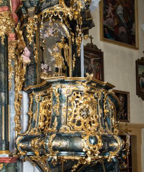 Zdjęcie nr 1: Ambona przyścienna, wisząca, ustawiona przy północnym filarze łuku tęczowego; dostępna jednobiegowymi schodami z balustradą od strony prezbiterium. Kosz na planie czworoboku, u dołu wydzielony silnie występującym ćwierćwałkiem z nałożoną złoconą dekoracją rocaille'ową; u góry zamknięty profilowanym gzymsem. Podwieszenie w formie odwróconego stożka. Ścianki kosza artykułowane pilastrami w kształcie spływów wolutowych. W polach prostokątne płyciny o wklęsłych narożach ozdobione bogato ażurowym ornamentem rokokowym. Zaplecek flankowany pilastrami podtrzymującymi przerwane belkowanie, które nad polem środkowym przechodzi w baldachim. W polu zaplecka prostokątna płycina zamknięta łukiem nadwieszonym z płaskorzeźbą św. Jana Nepomucena. Baldachim krótki, na rzucie półkolistym bez podniebienia, od dołu zdobiony lambrekinem. Na impostach cokoły utworzone z wolut, a na nich ustawione płomieniste wazony. Zwieńczenie w kształcie zbliżonym do kwadratu, ujętym z każdej strony spływami wolutowymi. W polu głównym owalna płycina, na której tle jest promienista gloria z gołębicą Ducha Świętego. Balustrada schodów pełna; na końcu zakończona filarkiem z rokokowym wazonem płomienistym. Całość silnie dekorowana ornamentem rocaille’owym. Struktura marmoryzowana w kolorze ciemnozielonym, ornamentyka i detale złocone oraz srebrzone.
