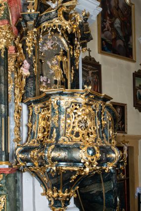 Zdjęcie nr 1: Ambona przyścienna, wisząca, ustawiona przy północnym filarze łuku tęczowego; dostępna jednobiegowymi schodami z balustradą od strony prezbiterium. Kosz na planie czworoboku, u dołu wydzielony silnie występującym ćwierćwałkiem z nałożoną złoconą dekoracją rocaille'ową; u góry zamknięty profilowanym gzymsem. Podwieszenie w formie odwróconego stożka. Ścianki kosza artykułowane pilastrami w kształcie spływów wolutowych. W polach prostokątne płyciny o wklęsłych narożach ozdobione bogato ażurowym ornamentem rokokowym. Zaplecek flankowany pilastrami podtrzymującymi przerwane belkowanie, które nad polem środkowym przechodzi w baldachim. W polu zaplecka prostokątna płycina zamknięta łukiem nadwieszonym z płaskorzeźbą św. Jana Nepomucena. Baldachim krótki, na rzucie półkolistym bez podniebienia, od dołu zdobiony lambrekinem. Na impostach cokoły utworzone z wolut, a na nich ustawione płomieniste wazony. Zwieńczenie w kształcie zbliżonym do kwadratu, ujętym z każdej strony spływami wolutowymi. W polu głównym owalna płycina, na której tle jest promienista gloria z gołębicą Ducha Świętego. Balustrada schodów pełna; na końcu zakończona filarkiem z rokokowym wazonem płomienistym. Całość silnie dekorowana ornamentem rocaille’owym. Struktura marmoryzowana w kolorze ciemnozielonym, ornamentyka i detale złocone oraz srebrzone.
