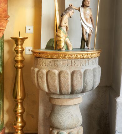 Zdjęcie nr 2: Kamienna chrzcielnica umieszczona na niewielkiej, kolistej stopie. Trzon gładki, w formie dwulalkowej tralki, przedzielonej pierścieniem z kolistymi guzami. Czara w kształcie półkuli, zdobiona motywem puklowania, powyżej inskrypcja: „NISI QUIS RENAT, FUERIT EX AQUA ET SPIRITUS SANCT”. Pokrywa drewniana na planie koła z grupą rzeźbiarską ukazującą scenę Chrztu Chrystusa; pokrywa na brzegu zdobiona pasem kimationu. Chrystus ukazany ze złożonymi rękami, stoi w wodzie, po jego lewej stronie stoi św. Jan Chrzciciel, który w lewej ręce trzyma wysoki krzyż, a prawą polewa wodą głowę Chrystusa. Zbawiciel ma spiczastą brodę i nos, duże oczy, włosy opadają na plecy, a nad czołem spływającą wodę. Sylwetki postaci szczupłe o sumarycznie oddanych detalach anatomicznych. Chrystus opasany w biodrach złotą tkaniną z zieloną podszewką, a św. Jan Chrzciciel w srebrzoną szatę przerzuconą przez prawe ramię i osłaniającą biodra. Za nimi ustawiona jest na długim promieniu gloria z gołębicą Ducha Świętego. Wewnątrz, na brzegu chrzcielnicy wyryta jest inskrypcja: „ANNO DNI 1746 DIEX DEC.”