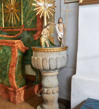 Zdjęcie nr 1: Kamienna chrzcielnica umieszczona na niewielkiej, kolistej stopie. Trzon gładki, w formie dwulalkowej tralki, przedzielonej pierścieniem z kolistymi guzami. Czara w kształcie półkuli, zdobiona motywem puklowania, powyżej inskrypcja: „NISI QUIS RENAT, FUERIT EX AQUA ET SPIRITUS SANCT”. Pokrywa drewniana na planie koła z grupą rzeźbiarską ukazującą scenę Chrztu Chrystusa; pokrywa na brzegu zdobiona pasem kimationu. Chrystus ukazany ze złożonymi rękami, stoi w wodzie, po jego lewej stronie stoi św. Jan Chrzciciel, który w lewej ręce trzyma wysoki krzyż, a prawą polewa wodą głowę Chrystusa. Zbawiciel ma spiczastą brodę i nos, duże oczy, włosy opadają na plecy, a nad czołem spływającą wodę. Sylwetki postaci szczupłe o sumarycznie oddanych detalach anatomicznych. Chrystus opasany w biodrach złotą tkaniną z zieloną podszewką, a św. Jan Chrzciciel w srebrzoną szatę przerzuconą przez prawe ramię i osłaniającą biodra. Za nimi ustawiona jest na długim promieniu gloria z gołębicą Ducha Świętego. Wewnątrz, na brzegu chrzcielnicy wyryta jest inskrypcja: „ANNO DNI 1746 DIEX DEC.”