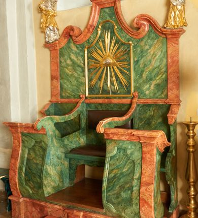 Zdjęcie nr 1: Ława na rzucie prostokąta,  ustawiona na cokole, dłuższym bokiem przylega do ściany. Trzy miejsca siedzące, oddzielone od siebie ściankami, środkowe najszersze. Centralne miejsce z wejściem na osi, boczne z wejściami od wschodu i zachodu, od przodu są zamknięte falisto-wypukłą ścianką. Naroża flankują pilastry, których kapitele tworzą gzyms, będący jednocześnie parapetem. Zaplecek oddzielony jest od dolnej części gzymsem. W jego centralnej części znajduje się wydzielone laskowaniem pole w kształcie prostokąta zbliżonego do kwadratu, zamkniętego łukiem o wklęsło-wypukłym kształcie utworzonym z wydłużonych wolut. W polu Oko Opatrzności na tle promienistej glorii. Zaplecek flankują pilastry, na których ustawione są rzeźby: po prawej św. Weroniki, a po lewej prawdopodobnie św. Marii Magdaleny. Pilastry przechodzą w falisty gzyms, wolutowo zawinięty do środka i zwężający się ku górze. Na poziomym odcinku korona zamknięta zakończona krzyżem maltańskim. Struktura polichromowana na kolor ciemnozielony; cokół, pilastry i gzymsy na kolor czerwony, detale złocone i srebrzone.