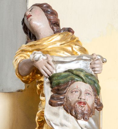 Zdjęcie nr 1: Rzeźba ścięta z tyłu przedstawiająca św. Weronikę. Święta ukazana w postawie stojącej o esowato wygiętej sylwetce z veraikonem w dłoni. Twarz świętej podłużna o ekspresyjnym wyrazie z otwartymi ustami i głową odchyloną do tyłu. Włosy bujne, długie, zasłaniające uszy i głębokimi falami opadające na plecy. Ubrana jest w długą, złoconą suknię z podwiniętymi rękawami, silnie drapowaną. Ma bose stopy. W dłoniach trzyma srebrzoną chustę z wizerunkiem Chrystusa. Twarz Zbawiciela pełna, okolona średniej długości brodą i bujnymi włosami rozwianymi na boki. Na głowie ma szeroką, zieloną koronę cierniową. Polichromia w odsłoniętych partiach ciała naturalistyczna. 