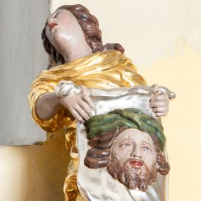 Zdjęcie nr 1: Rzeźba ścięta z tyłu przedstawiająca św. Weronikę. Święta ukazana w postawie stojącej o esowato wygiętej sylwetce z veraikonem w dłoni. Twarz świętej podłużna o ekspresyjnym wyrazie z otwartymi ustami i głową odchyloną do tyłu. Włosy bujne, długie, zasłaniające uszy i głębokimi falami opadające na plecy. Ubrana jest w długą, złoconą suknię z podwiniętymi rękawami, silnie drapowaną. Ma bose stopy. W dłoniach trzyma srebrzoną chustę z wizerunkiem Chrystusa. Twarz Zbawiciela pełna, okolona średniej długości brodą i bujnymi włosami rozwianymi na boki. Na głowie ma szeroką, zieloną koronę cierniową. Polichromia w odsłoniętych partiach ciała naturalistyczna. 