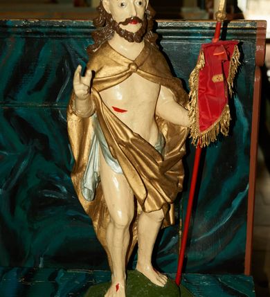 Zdjęcie nr 1: Rzeźba Chrystusa Zmartwychwstałego pełnoplastyczna, wolnostojąca. Chrystus ukazany jest w całej postaci, ustawiony frontalnie, w delikatnym kontrapoście; prawą, wysoko uniesioną ręką czyni gest błogosławieństwa, a w lewej trzyma chorągiew o długim drzewcu zakończonym krzyżem, na czerwonej tkaninie chorągwi widnieje złoty krzyż. Chrystus ma podłużną twarz z długim i wąskim nosem, wzrokiem skierowanym w dal oraz krótką i ciemną brodę, rozdzieloną na końcu w dwa pukle. Włosy długie i ciemnobrązowe, opadają na plecy i ramiona. Chrystus jest ubrany w złocony płaszcz spięty na piersi, z lewą połą przerzuconą przez prawą rękę. Na biodrach ma założone srebrzone perizonium. Na dłoniach, stopach i boku zaznaczone są ślady męki. Polichromia ciała jest naturalistyczna. Rzeźba ustawiona jest na niskiej podstawie w kształcie prostokąta o ściętych narożach, na którym osadzona jest zielona skała.
