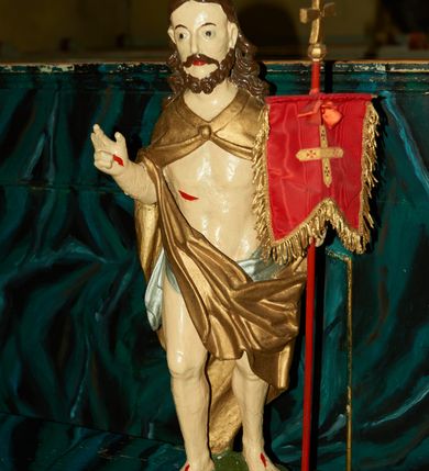 Zdjęcie nr 2: Rzeźba Chrystusa Zmartwychwstałego pełnoplastyczna, wolnostojąca. Chrystus ukazany jest w całej postaci, ustawiony frontalnie, w delikatnym kontrapoście; prawą, wysoko uniesioną ręką czyni gest błogosławieństwa, a w lewej trzyma chorągiew o długim drzewcu zakończonym krzyżem, na czerwonej tkaninie chorągwi widnieje złoty krzyż. Chrystus ma podłużną twarz z długim i wąskim nosem, wzrokiem skierowanym w dal oraz krótką i ciemną brodę, rozdzieloną na końcu w dwa pukle. Włosy długie i ciemnobrązowe, opadają na plecy i ramiona. Chrystus jest ubrany w złocony płaszcz spięty na piersi, z lewą połą przerzuconą przez prawą rękę. Na biodrach ma założone srebrzone perizonium. Na dłoniach, stopach i boku zaznaczone są ślady męki. Polichromia ciała jest naturalistyczna. Rzeźba ustawiona jest na niskiej podstawie w kształcie prostokąta o ściętych narożach, na którym osadzona jest zielona skała.