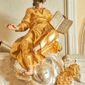 Zdjęcie nr 1: Rzeźba św. Marka, który siedzi na srebrnych obłokach. Święty jest ukazany frontalnie, delikatnie wygięty do tyłu z głową uniesioną do góry w prawą stronę. Nogi ma ugięte w kolanach, lewą uniesioną i odwiedzioną na bok; ręce szeroko rozłożone na boki, w lewej dłoni trzyma otwartą księgę. Twarz szeroka z dużym i szerokim na końcu nosem, dużymi oczami oraz silnie zarumienionymi policzkami. Włosy krótkie, ciemnobrązowe z zakolami nad czołem, broda krótka i postrzępiona, ułożona w dwa duże pukle. Święty ubrany jest w długą i złoconą suknię, drapowaną w grube i ostro łamane fałdy z dekoltem w kształcie litery „V”. Po lewej stronie świętego znajduje się złocone popiersie lwa. Polichromia naturalistyczna w odsłoniętych partiach ciała świętego, szaty i lew złocone, obłoki srebrzone. 
