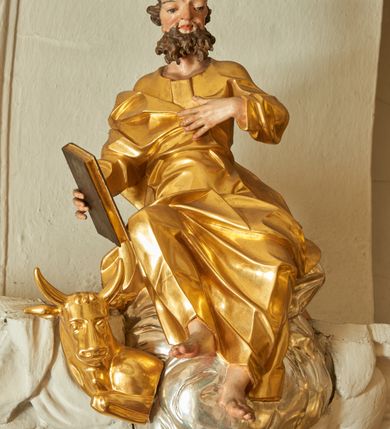 Zdjęcie nr 3: Rzeźba św. Łukasza, który siedzi na srebrnych obłokach. Święty jest ukazany frontalnie, w prawej dłoni trzyma zamkniętą księgę, a lewą dłoń składa na piersi, nogi ma ugięte w kolanach, prawą delikatnie uniesioną do góry. Twarz szeroka z dużym i szerokim na końcu nosem, dużymi oczami oraz silnie zarumienionymi policzkami. Włosy krótkie, ciemnobrązowe z łysiną nad czołem, broda krótka i postrzępiona, ułożona w dwa duże pukle. Święty ubrany jest w długą i złoconą suknię, drapowaną w grube i ostro łamane fałdy. Po prawej stronie świętego znajduje się złocone popiersie wołu. Polichromia naturalistyczna w odsłoniętych partiach ciała świętego, szaty i wół złocone, obłoki srebrzone. 
