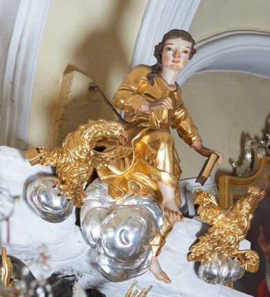 Zdjęcie nr 1: Rzeźba św. Jana, który siedzi na srebrnych obłokach z prawą nogą ugiętą w kolanie i uniesioną lekko do góry. Święty jest delikatnie zwrócony w prawo z głową skierowaną w prawą stronę, w lewej wyprostowanej  ręce trzyma zamkniętą księgę, a prawą rękę ma ugiętą w łokciu. Twarz szeroka z dużym i szerokim na końcu nosem, dużymi oczami oraz silnie zarumienionymi policzkami i nosem. Włosy długie, ciemnobrązowe opadają na prawe ramię. Święty ubrany jest w długą, złoconą suknię z kołnierzykiem u szyi, drapowaną w grube i ostro łamane fałdy. Po bokach świętego siedzą dwa orły, które trzymają w szponach kałamarze. Polichromia naturalistyczna w odsłoniętych partiach ciała, szaty i orły złocone, obłoki srebrzone.