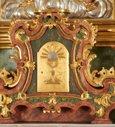 Zdjęcie nr 1: Tabernakulum w bogatej ornamentalnej oprawie, ustawione na niskim cokole. Obramienie tabernakulum utworzone z połączonych ze sobą wolut, zwężające się ku górze, bogato dekorowane ornamentem rocaille’owym. W zwieńczeniu niska podstawa służąca jako tron wystawienia. W polu głównym płycina w kształcie stojącego prostokąta o wklęsłych bokach, zamknięta łukiem wklęsło-wypukłym. Drzwiczki przyjmują kształt stojącego prostokąta zamkniętego łukiem półkolistym nadwieszonym; w polu płaskorzeźbione przedstawienie kielicha z hostią w glorii na gładkim tle. Struktura marmoryzowana w kolorze ciemnozielonym, profilowania na czerwono, ornamentyka złocona. 

