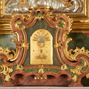 Zdjęcie nr 1: Tabernakulum w bogatej ornamentalnej oprawie, ustawione na niskim cokole. Obramienie tabernakulum utworzone z połączonych ze sobą wolut, zwężające się ku górze, bogato dekorowane ornamentem rocaille’owym. W zwieńczeniu niska podstawa służąca jako tron wystawienia. W polu głównym płycina w kształcie stojącego prostokąta o wklęsłych bokach, zamknięta łukiem wklęsło-wypukłym. Drzwiczki przyjmują kształt stojącego prostokąta zamkniętego łukiem półkolistym nadwieszonym; w polu płaskorzeźbione przedstawienie kielicha z hostią w glorii na gładkim tle. Struktura marmoryzowana w kolorze ciemnozielonym, profilowania na czerwono, ornamentyka złocona. 
