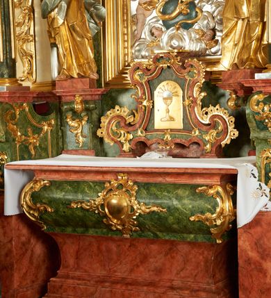 Zdjęcie nr 2: Tabernakulum w bogatej ornamentalnej oprawie, ustawione na niskim cokole. Obramienie tabernakulum utworzone z połączonych ze sobą wolut, zwężające się ku górze, bogato dekorowane ornamentem rocaille’owym. W zwieńczeniu niska podstawa służąca jako tron wystawienia. W polu głównym płycina w kształcie stojącego prostokąta o wklęsłych bokach, zamknięta łukiem wklęsło-wypukłym. Drzwiczki przyjmują kształt stojącego prostokąta zamkniętego łukiem półkolistym nadwieszonym; w polu płaskorzeźbione przedstawienie kielicha z hostią w glorii na gładkim tle. Struktura marmoryzowana w kolorze ciemnozielonym, profilowania na czerwono, ornamentyka złocona. 
