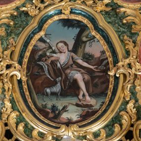 Zdjęcie nr 1: Obraz w kształcie owalu o wybrzuszonej górnej części z przedstawieniem młodzieńczego św. Jana Chrzciciela. Święty został ukazany w centrum kompozycji w pozycji siedzącej, odchylony do tyłu. Prawą stopę ma wysuniętą do przodu i wspiera ją na kamieniu. W lewej dłoni święty trzyma długi krzyż obwiedziony białą banderolą z napisem: „ECCE AGNUS DEI,”  a lewą wyciąga w bok i nabiera w muszlę wody z wytryskającego z boku obrazu źródełka. Święty ma owalną twarz z wysokim czołem, długim nosem, małymi ustami oraz przymrużonymi oczami. Jego długie i bujne włosy spływają na plecy. Karnacja ciała jasna, miejscami silnie zarumieniona. Święty ubrany jest w krótką, brązową skórę wielbłądzią, przewiązaną w talii czarnym paskiem, odsłaniającym prawe nagie ramię i tors. Po lewej stronie obrazu stoi zwrócony w stronę św. Jana mały, biały baranek. Kompozycja ukazana jest na tle bujnej roślinności i skalnego krajobrazu. Pośrodku kompozycji za plecami świętego stoi wysokie drzewo. W oddali widoczne jest różowo-niebieskie niebo. 