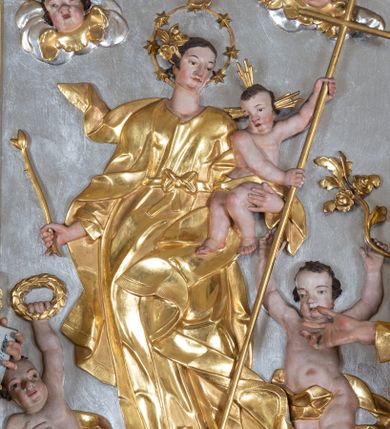 Zdjęcie nr 1: Płaskorzeźba w kształcie prostokąta zamkniętego łukiem wklęsło-wypukłym zdobionym grzebieniami rocaille’owymi pośrodku. W polu, na jednolicie srebrnym tle ukazana jest Matka Boska Niepokalanie Poczęta. Maria stoi na globie ziemskim, prawą stopą depcze węża, a lewą trzyma na półksiężycu. Jej sylwetka jest esowato wygięta, ma długą szyję. Matka Boska w lewej dłoni trzyma Dzieciątko Jezus, a w prawej berło. Twarz szeroka z nienaturalnie dużym nosem, wąskimi ustami i silnie zarumienionymi policzkami. Włosy długie, ciemnobrązowe, spięte do tyłu i ozdobione płaskorzeźbionymi różami. Maria ubrana jest w długą suknię, przewiązaną w pasie wstążką związaną na kokardkę oraz złocony płaszcz, na stopach ma sandały, a wokół głowy okrągły nimb z siedmioma gwiazdami. Dzieciątko ukazane jest w pozycji siedzącej, po swojej lewej stronie trzyma w dłoniach długi krzyż, którego drzewiec wbija się w węża. Twarz Dzieciątka ma nienaturalny gruszkowaty kształt z dużym nosem i wyraźnie wydatnym podbródkiem, policzki silnie zarumienione. Włosy krótkie, ciemnobrązowe, wokół głowy promienisty nimb krzyżowy. Dzieciątko ubrane jest w złoconą przepaskę biodrową. Glob ziemski w kolorze niebieskim osadzony na chmurze srebrzonych obłoków, z której wyłaniają się dwie uskrzydlone główki anielskie. Wyżej, dwa aniołki unoszące do góry ręce. Aniołek po lewej stronie trzyma w dłoni wieniec chwały, a aniołek po prawej gałązkę róży. W górnej części obrazu, kolejne dwie uskrzydlone główki anielskie. Polichromia w odsłoniętych partiach ciała naturalistyczna, szaty i atrybuty złocone, detale srebrzone.