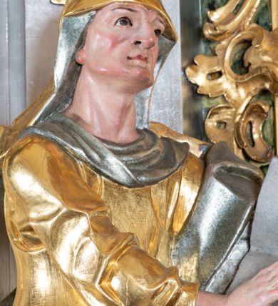 Zdjęcie nr 7: Rzeźba św. Anny ścięta z tyłu, drążona, ustawiona na prostopadłościennym, marmoryzowanym na jasnoczerwono cokole. Święta ukazana jest frontalnie w silnym kontrapoście z głową uniesioną ku górze. Ręce ma skierowane na lewy bok, trzyma w nich otwartą księgę zapisaną słowami: „Merito igitur / germen eadem / Anaa divino germo reult / Spiritus pierna liberi libent / Labori qve durionustructa / Vertat am sancti ludaribusmais / Clamorite me cum opta / veram”. Twarz szeroka o wyraźnie zaznaczonych kościach policzkowych, dużych oczach, małych ustach i nienaturalnie dużym nosie, policzki silnie zarumienione. Święta ubrana jest w długą suknię z długimi i wąskimi rękawami i gorsetem, drapowaną w grube fałdy. Na głowę ma nałożony złocony welon ze srebrzoną podszewką, a na lewe ramię złocony płaszcz ze srebrzoną podszewką, który zawija się z prawej strony ciała. Polichromia naturalistyczna w odsłoniętych partiach ciała; szaty i atrybuty złocone i srebrzone.
