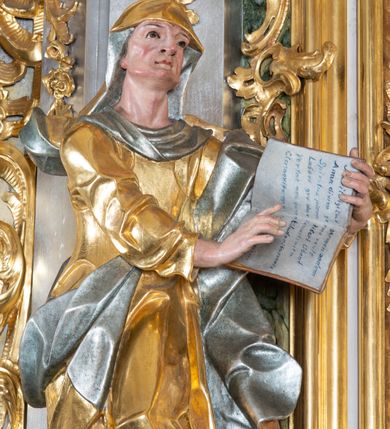 Zdjęcie nr 4: Rzeźba św. Anny ścięta z tyłu, drążona, ustawiona na prostopadłościennym, marmoryzowanym na jasnoczerwono cokole. Święta ukazana jest frontalnie w silnym kontrapoście z głową uniesioną ku górze. Ręce ma skierowane na lewy bok, trzyma w nich otwartą księgę zapisaną słowami: „Merito igitur / germen eadem / Anaa divino germo reult / Spiritus pierna liberi libent / Labori qve durionustructa / Vertat am sancti ludaribusmais / Clamorite me cum opta / veram”. Twarz szeroka o wyraźnie zaznaczonych kościach policzkowych, dużych oczach, małych ustach i nienaturalnie dużym nosie, policzki silnie zarumienione. Święta ubrana jest w długą suknię z długimi i wąskimi rękawami i gorsetem, drapowaną w grube fałdy. Na głowę ma nałożony złocony welon ze srebrzoną podszewką, a na lewe ramię złocony płaszcz ze srebrzoną podszewką, który zawija się z prawej strony ciała. Polichromia naturalistyczna w odsłoniętych partiach ciała; szaty i atrybuty złocone i srebrzone.
