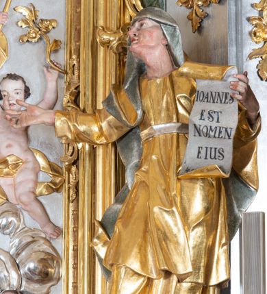 Zdjęcie nr 1: Rzeźba św. Elżbiety ścięta z tyłu, drążona, ustawiona na prostopadłościennym, marmoryzowanym na jasnoczerwono cokole. Święta jest ukazana frontalnie w silnym kontrapoście z głową uniesioną ku górze. Ręce ma rozłożone szeroko na boki, w lewej trzyma kartę z napisem: „IOANNES / EST / NOMEN / EIUS / Ann(?)”. Twarz szeroka o wyraźnie zaznaczonych kościach policzkowych, dużych oczach, małych ustach i nienaturalnie dużym nosie, policzki silnie zarumienione. Święta ubrana jest w długą suknię z długimi i wąskimi rękawami, przewiązaną w talli paskiem, drapowaną w grube i ostro łamane fałdy. Na głowę ma nałożony srebrzony welon. Polichromia naturalistyczna w odsłoniętych partiach ciała; szaty i atrybuty złocone i srebrzone.
