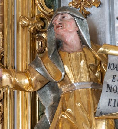 Zdjęcie nr 2: Rzeźba św. Elżbiety ścięta z tyłu, drążona, ustawiona na prostopadłościennym, marmoryzowanym na jasnoczerwono cokole. Święta jest ukazana frontalnie w silnym kontrapoście z głową uniesioną ku górze. Ręce ma rozłożone szeroko na boki, w lewej trzyma kartę z napisem: „IOANNES / EST / NOMEN / EIUS / Ann(?)”. Twarz szeroka o wyraźnie zaznaczonych kościach policzkowych, dużych oczach, małych ustach i nienaturalnie dużym nosie, policzki silnie zarumienione. Święta ubrana jest w długą suknię z długimi i wąskimi rękawami, przewiązaną w talli paskiem, drapowaną w grube i ostro łamane fałdy. Na głowę ma nałożony srebrzony welon. Polichromia naturalistyczna w odsłoniętych partiach ciała; szaty i atrybuty złocone i srebrzone.
