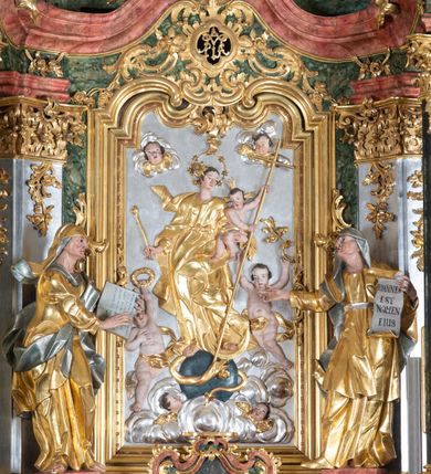 Zdjęcie nr 4: Rzeźba św. Elżbiety ścięta z tyłu, drążona, ustawiona na prostopadłościennym, marmoryzowanym na jasnoczerwono cokole. Święta jest ukazana frontalnie w silnym kontrapoście z głową uniesioną ku górze. Ręce ma rozłożone szeroko na boki, w lewej trzyma kartę z napisem: „IOANNES / EST / NOMEN / EIUS / Ann(?)”. Twarz szeroka o wyraźnie zaznaczonych kościach policzkowych, dużych oczach, małych ustach i nienaturalnie dużym nosie, policzki silnie zarumienione. Święta ubrana jest w długą suknię z długimi i wąskimi rękawami, przewiązaną w talli paskiem, drapowaną w grube i ostro łamane fałdy. Na głowę ma nałożony srebrzony welon. Polichromia naturalistyczna w odsłoniętych partiach ciała; szaty i atrybuty złocone i srebrzone.
