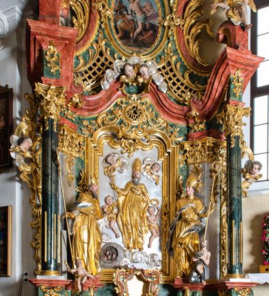 Zdjęcie nr 2: Ołtarz przyścienny, architektoniczny z mensą w formie sarkofagu o wklęsło-wypukłych bokach. W mensie kamienny portatyl. Retabulum jednoosiowe, jednokondygnacyjne ze zwieńczeniem, ustawione na wysokim zdwojonym cokole, zryzalitowanym bo bokach. Nastawa ujęta parą narożnych pilastrów kompozytowych oraz parą kolumn znacznie wysuniętych przed lico ołtarza. Kolumny o kanelowanych trzonach i kompozytowych głowicach podtrzymują faliste belkowanie. Zwieńczenie ujęte po bokach motywami „S” kształtnymi o wolutowo zwiniętych końcach, z których dolne mocno wysunięte do przodu, spoczywają na belkowaniu. Zwieńczenie zamknięte dwoma stykającymi się wolutami. Po bokach retabulum ażurowe uszaki formowane z grzebieni rocaille&#039;owych i motywów „C” kształtnych z rzeźbioną, uskrzydloną główką anielską. 
Pole środkowe w kształcie prostokątnej wnęki z zamknięciem kształtowanym z motywów „C”. We wnęce płaskorzeźba z przedstwieniem św. Mikołaja ujęta podwójną profilowaną ramą. U dołu, po bokach wnęki dwie konsole z ustawionymi pełnoplastycznymi figurami św. Augustyna po lewej oraz św. Marcina (?) po prawej stronie. Ponad kolumnami, po bokach zwieńczenia dwie figury aniołów, powyżej para uskrzydlonych puttów. W zwieńczeniu owalna wnęka z obrazem przedstawiającym św. Sebastiana, ujętym podwójną ramą. 
Ołtarz bogato dekorowany. Mensa zdobiona ornamentem rocaille&#039;owym. Pola cokołów zdobione płycinami kształtowanymi z grzebieni rocaille&#039;owych dekorujących też fryz. Trzony kolumn i pilastrów zdobią podwieszane motywy rocaille&#039;owe przeplatane z gałązkami róż, tworzącymi również rozległy, ażurowy kartusz nad polem środkowym. Boki zwieńczenia ujęte ażurowymi liśćmi akantu. Poniżej wnęki ażurowa, ukośna kratka regencyjna. Boki wnęki i szczyt zwieńczenia dekorowane pełnoplastycznymi rocaille&#039;ami. 
Całość struktury ołtarza pokryta malowaną marmoryzacją w odcieniach zieleni i brązu. Detal architektoniczny złocony, pilastry srebrzone.