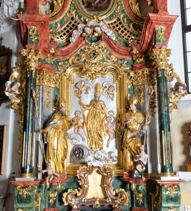 Zdjęcie nr 1: Ołtarz przyścienny, architektoniczny z mensą w formie sarkofagu o wklęsło-wypukłych bokach. W mensie kamienny portatyl. Retabulum jednoosiowe, jednokondygnacyjne ze zwieńczeniem, ustawione na wysokim zdwojonym cokole, zryzalitowanym bo bokach. Nastawa ujęta parą narożnych pilastrów kompozytowych oraz parą kolumn znacznie wysuniętych przed lico ołtarza. Kolumny o kanelowanych trzonach i kompozytowych głowicach podtrzymują faliste belkowanie. Zwieńczenie ujęte po bokach motywami „S” kształtnymi o wolutowo zwiniętych końcach, z których dolne mocno wysunięte do przodu, spoczywają na belkowaniu. Zwieńczenie zamknięte dwoma stykającymi się wolutami. Po bokach retabulum ażurowe uszaki formowane z grzebieni rocaille'owych i motywów „C” kształtnych z rzeźbioną, uskrzydloną główką anielską. 
Pole środkowe w kształcie prostokątnej wnęki z zamknięciem kształtowanym z motywów „C”. We wnęce płaskorzeźba z przedstwieniem św. Mikołaja ujęta podwójną profilowaną ramą. U dołu, po bokach wnęki dwie konsole z ustawionymi pełnoplastycznymi figurami św. Augustyna po lewej oraz św. Marcina (?) po prawej stronie. Ponad kolumnami, po bokach zwieńczenia dwie figury aniołów, powyżej para uskrzydlonych puttów. W zwieńczeniu owalna wnęka z obrazem przedstawiającym św. Sebastiana, ujętym podwójną ramą. 
Ołtarz bogato dekorowany. Mensa zdobiona ornamentem rocaille'owym. Pola cokołów zdobione płycinami kształtowanymi z grzebieni rocaille'owych dekorujących też fryz. Trzony kolumn i pilastrów zdobią podwieszane motywy rocaille'owe przeplatane z gałązkami róż, tworzącymi również rozległy, ażurowy kartusz nad polem środkowym. Boki zwieńczenia ujęte ażurowymi liśćmi akantu. Poniżej wnęki ażurowa, ukośna kratka regencyjna. Boki wnęki i szczyt zwieńczenia dekorowane pełnoplastycznymi rocaille'ami. 
Całość struktury ołtarza pokryta malowaną marmoryzacją w odcieniach zieleni i brązu. Detal architektoniczny złocony, pilastry srebrzone.