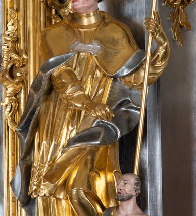 Zdjęcie nr 31: Ołtarz przyścienny, architektoniczny z mensą w formie sarkofagu o wklęsło-wypukłych bokach. W mensie kamienny portatyl. Retabulum jednoosiowe, jednokondygnacyjne ze zwieńczeniem, ustawione na wysokim zdwojonym cokole, zryzalitowanym bo bokach. Nastawa ujęta parą narożnych pilastrów kompozytowych oraz parą kolumn znacznie wysuniętych przed lico ołtarza. Kolumny o kanelowanych trzonach i kompozytowych głowicach podtrzymują faliste belkowanie. Zwieńczenie ujęte po bokach motywami „S” kształtnymi o wolutowo zwiniętych końcach, z których dolne mocno wysunięte do przodu, spoczywają na belkowaniu. Zwieńczenie zamknięte dwoma stykającymi się wolutami. Po bokach retabulum ażurowe uszaki formowane z grzebieni rocaille'owych i motywów „C” kształtnych z rzeźbioną, uskrzydloną główką anielską. 
Pole środkowe w kształcie prostokątnej wnęki z zamknięciem kształtowanym z motywów „C”. We wnęce płaskorzeźba z przedstwieniem św. Mikołaja ujęta podwójną profilowaną ramą. U dołu, po bokach wnęki dwie konsole z ustawionymi pełnoplastycznymi figurami św. Augustyna po lewej oraz św. Marcina (?) po prawej stronie. Ponad kolumnami, po bokach zwieńczenia dwie figury aniołów, powyżej para uskrzydlonych puttów. W zwieńczeniu owalna wnęka z obrazem przedstawiającym św. Sebastiana, ujętym podwójną ramą. 
Ołtarz bogato dekorowany. Mensa zdobiona ornamentem rocaille'owym. Pola cokołów zdobione płycinami kształtowanymi z grzebieni rocaille'owych dekorujących też fryz. Trzony kolumn i pilastrów zdobią podwieszane motywy rocaille'owe przeplatane z gałązkami róż, tworzącymi również rozległy, ażurowy kartusz nad polem środkowym. Boki zwieńczenia ujęte ażurowymi liśćmi akantu. Poniżej wnęki ażurowa, ukośna kratka regencyjna. Boki wnęki i szczyt zwieńczenia dekorowane pełnoplastycznymi rocaille'ami. 
Całość struktury ołtarza pokryta malowaną marmoryzacją w odcieniach zieleni i brązu. Detal architektoniczny złocony, pilastry srebrzone.